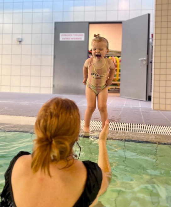 Een lachend kind staat aan de rand van het zwembad, terwijl een volwassene in het water het kind toespreekt. (vergroot foto)