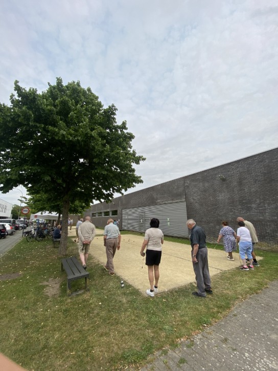 Groep speelt jeu de boules op een zandbaan naast een gebouw onder bewolkte lucht. (vergroot foto)