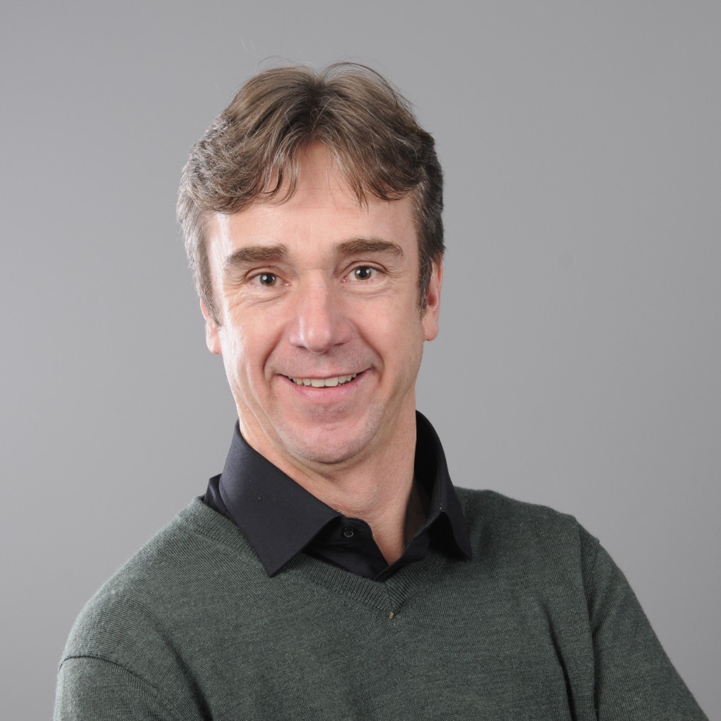 Portret van een lachende man met lichtbruin haar, gekleed in een donkergroene trui over een zwarte overhemd, voor een effen grijze achtergrond. (vergroot foto)