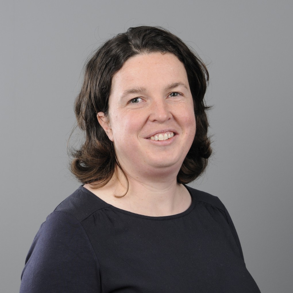 Portret van een vrouw met halflang donkerbruin haar, glimlachend, gekleed in een donkerblauw shirt, tegen een effen grijze achtergrond. (vergroot foto)