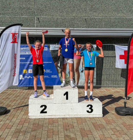 Drie meisjes met medailles staan op een podium, juichend bij een sportevenement in de zon. (vergroot foto)