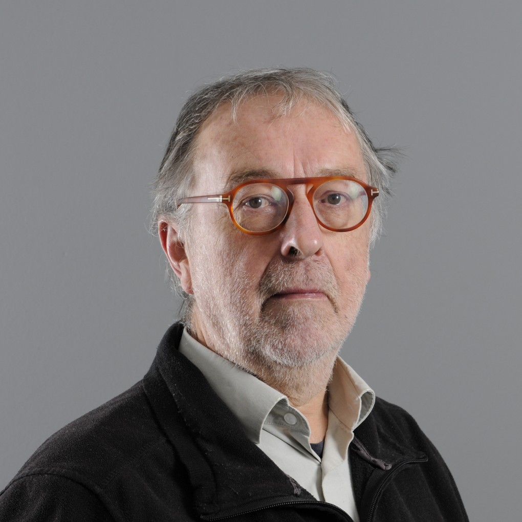 Portret van een oudere man met grijs haar en baard, die een lichtgrijs overhemd en een zwarte jas draagt. Hij heeft een serieuze blik en draagt een bril met een bruin/oranje montuur. De achtergrond is effen grijs. (vergroot foto)