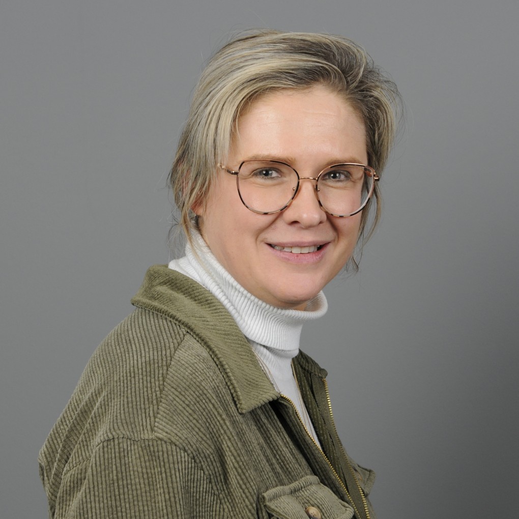 Portret van een vrouw met blond haar, die een groene corduroy jas en een witte coltrui draagt, met een lichte glimlach en een grijze achtergrond. (vergroot foto)