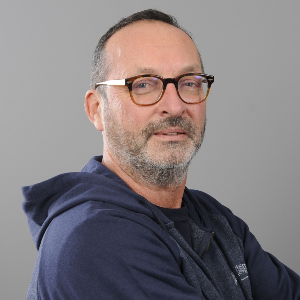 Portret van een man met kort grijs haar en baard, die een bril met bruin montuur draagt. Hij draagt een donkerblauwe hoodie en kijkt ontspannen naar de camera tegen een effen grijze achtergrond. (vergroot foto)