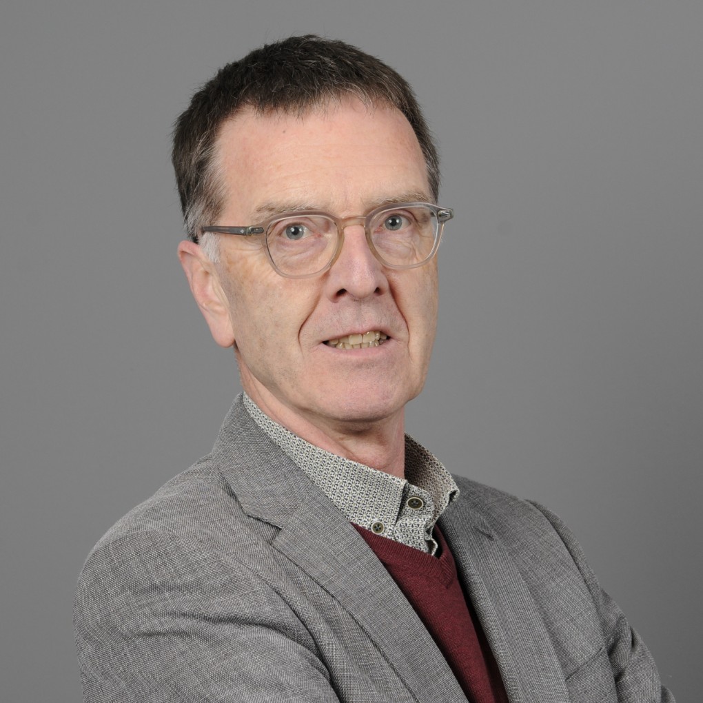 Portret van een oudere man met kort bruin haar en een grijze blazer, een roodbruine trui en een lichtgekleurde overhemd met patroon, tegen een grijze achtergrond. Hij draagt een bril en kijkt recht in de camera met een neutrale uitdrukking. (vergroot foto)